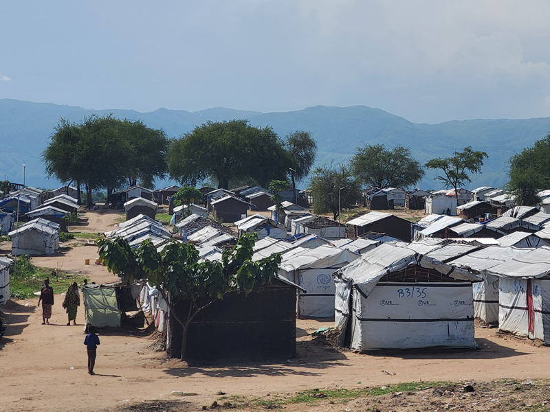 Young child in blue shirt collecting water from a community well, surrounded by people in colorful clothing at a rural settlement with wooden structures and dried earth ground, representing APROSHAV's commitment to accessing clean water and improving human health in underserved communities