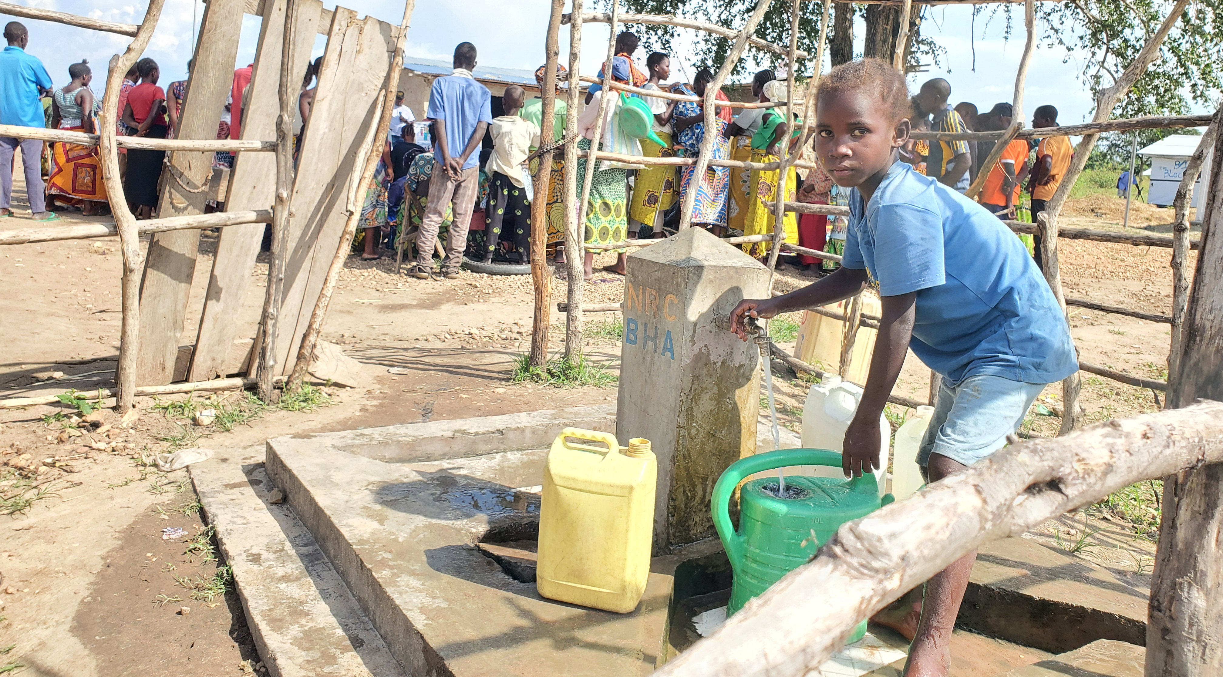 Young child in blue shirt collecting water from a community well, surrounded by people in colorful clothing at a rural settlement with wooden structures and dried earth ground, representing APROSHAV's commitment to accessing clean water and improving human health in underserved communities